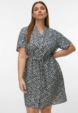 Hosszú barna hajú nő, rövid ujjú, gombos ruha visel, melynek sötétkék leveles mintája és derékkötője van, egyik kezét a csípőjén tartja.