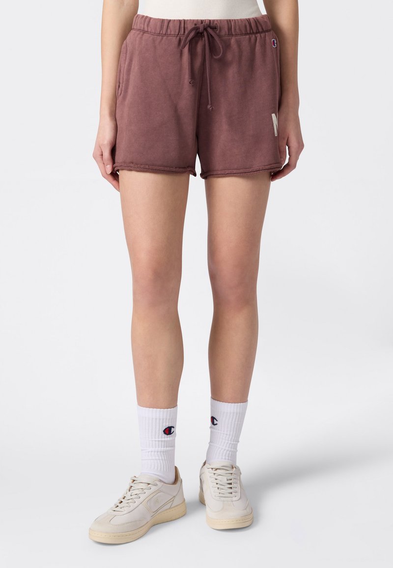 Person trägt braune Shorts mit Kordelzug, weiße Socken mit Champion-Logo und beige Turnschuhe und steht vor einem schlichten hellen Hintergrund.