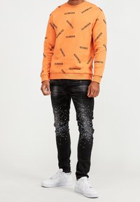 Sudadera naranja con "GLORIOUS" impreso en negro, combinada con jeans negros con un patrón de salpicaduras blancas y zapatillas deportivas blancas.