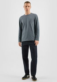 Langärmliges graues Baumwollhemd mit rundem Halsausschnitt, kombiniert mit dunklen Jeans und schwarzen Sneakers mit weißen und beigen Akzenten.