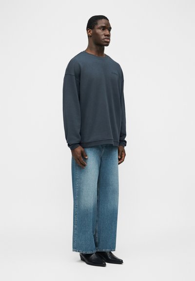 Άνδρας που στέκεται φορώντας έναν άνετο σκούρο γκρι φούτερ, φαρδιά μπλε τζιν και μαύρες μπότες με μυτερή μύτη, μπροστά σε σκηνικό απλού λευκού φόντου.