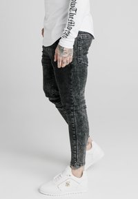 Vitt långärmad skjorta med svart tryckt text, kombinerad med mörkgrå slim-fit jeans och vita sneakers med en guldfärgad logotypdetalj.