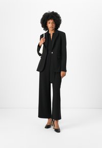 Zwarte op maat gemaakte blazer met één knoop, gecombineerd met wijde broekspijpen. Gladde stofstructuur, afgewerkt met zwarte hoge hakken en een klein wit tasje.