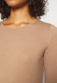 Maglione a coste marrone chiaro con scollo rotondo, maniche lunghe aderenti e dettagli delle cuciture sottili sulle spalle.