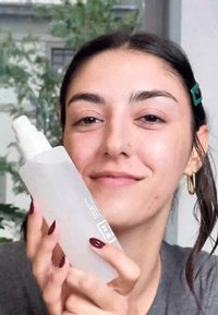 Frostad glas sprayflaska med en vit pump topp, hålls av en person med mörkt hår. Naglarna är djupt röda. Bakgrunden visar grön denna.