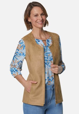 Beige Kunstlederweste mit Rundhalsausschnitt und Reißverschluss vorne, ausgestattet mit zwei Seitentaschen. Getragen über einem blauen Blümchen-Oberteil mit 3/4-Ärmeln.