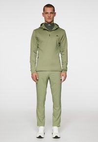 Vihreä urheiluasu, jossa on pitkät hihat ja vetoketjukaulus, yhdistettynä matching-housuihin ja valkoisiin lenkkareihin. Sisältää logo-yksityiskohtia.