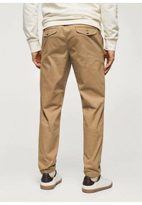Pantalon en coton beige à coupe décontractée, doté de deux poches arrière avec fermetures à bouton, associé à des baskets blanches et marron.