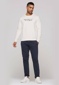 Men's light cream sweatshirt with printed text, paired with dark navy pants. Model wears white sneakers. Simple, casual design.