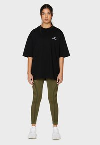 Sort oversized t-shirt med et lille logo, parret med olivengrønne leggings med netdetaljer og teksturerede mønstre. Hvide sneakers fuldender looket.
