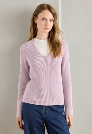 Jasnoróżowy sweter dzianinowy z dekoltem w kształcie litery V, o prążkowanej strukturze, dopasowanej sylwetce i długich rękawach, noszony na białej koszuli i zestawiony z jeansami.
