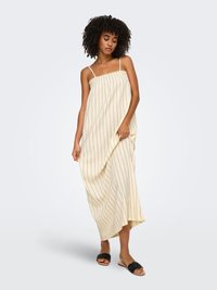 Femme aux cheveux bouclés portant une robe maxi sans manches crème à rayures et des sandales noires slip-on, debout sur un fond clair.