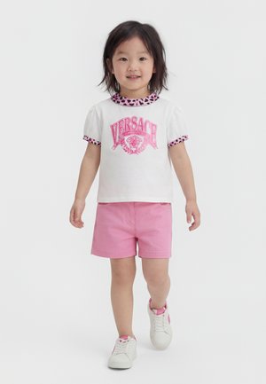 Jeune fille marche en avant portant un t-shirt blanc Versace avec un liseré léopard rose, un short rose et des baskets blanches avec des détails roses.