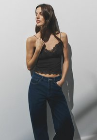 Melna mežģīņu apmaite ar plānām lencītēm, valkāta kopā ar tumšām džinsa biksēm ar augstu jostasvietu. Modele pozē gaišā fonā.