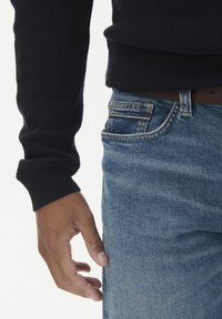 Kairioje ranka atsipalaidavusi šalia mėlynų džinsų su ruduo diržu ir juodo megztinio rankovė matoma prieš baltą foną.