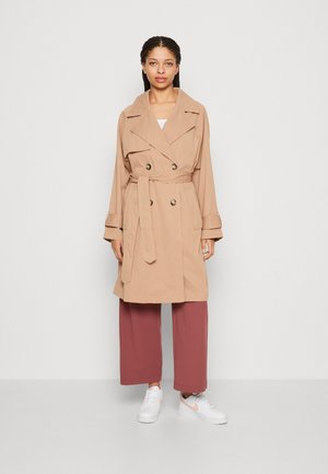 Manteau en trench beige avec des boutons double-breasted, un large col et une ceinture. Porté par-dessus un pantalon large bordeaux et des baskets blanches.