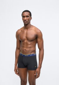 Homme musclé portant un boxer noir Nike avec une ceinture bleue et jaune, debout devant un fond blanc uni.