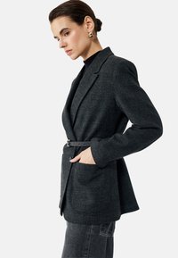 Grijze geruite blazer met een getailleerde taille, lange mouwen en zijzakken. Beschikt over een contrasterende riem, gemaakt van textuurstof.