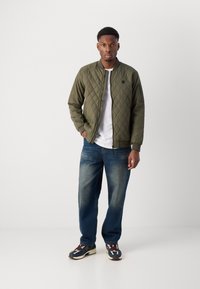 Veste bomber matelassée vert olive avec un col côtelé, portée sur une chemise blanche, associée à un jean bleu et des baskets multicolores.