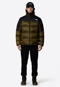 Oliwkowa puchowa kurtka z czarnymi ramionami, wysokim kołnierzem i zapięciem na zamek. Połączona z czarnymi spodniami i brązowymi butami turystycznymi, noszona z czarną czapką.