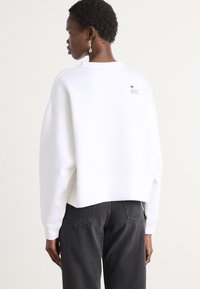 White cropped sweatshirt with a round neckline and dropped shoulders. Features a small black Lacoste logo label on the back. Paired with dark gray jeans.