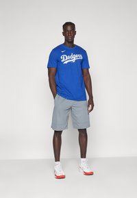 Maglietta in cotone blu con logo "Dodgers" in bianco, abbinata a pantaloni corti grigi. Le scarpe sono bianche con dettagli rossi. Un abbigliamento sportivo semplice e informale.