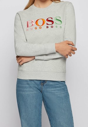 Person wearing light grey sweatshirt with colorful "BOSS HUGO BOSS" logo embroidery across chest and blue denim jeans, arms crossed.