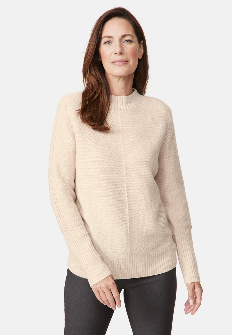 Beige Langarmpullover mit einem gerippten Ausschnitt und Bündchen, der ein strukturiertes Strickdesign und eine gerade Passform aufweist. Getragen mit dunkelgrauen Hosen.