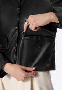 Giacca di pelle nera con bottoni dorati, tasca frontale e una leggera lucentezza. La mano del modello è posizionata sulla giacca, evidenziando il dettaglio della tasca.