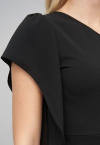 Robe noire avec un corsage ajusté et des manches évasées à la texture lisse. Des coutures remarquables le long de l'ourlet et des épaules rehaussent le design.