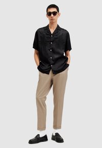 Camicia nera a maniche corte con finitura satinata e design ricamato di un'aquila, abbinata a pantaloni beige e mocassini neri con calzini bianchi.