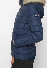 Mörkblå vadderad jacka med pälskantad huva, sidofickor och liten flagglogotyp på bröstet och ärmen, bärs av en person i svarta byxor.