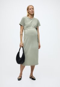 Έγκυος γυναίκα που φοράει φόρεμα μήκους μίντι με πράσινες και λευκές ρίγες, κρατώντας μια μαύρη τσάντα και φορώντας μαύρα παπούτσια Mary Jane, σε φόντο λευκού.