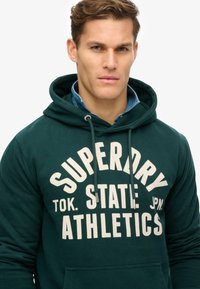 Sudadera verde oscuro con texto impreso en blanco "SUPERDRY STATE TOK. JPN. ATHLETICS." Capucha con cordón y bolsillo tipo canguro en la parte delantera, material suave.