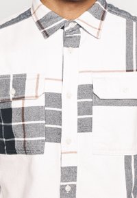 Camicia bianca con pattern a quadri neri e grigi e dettagli marroni. Presenta un colletto standard e tasche frontali.