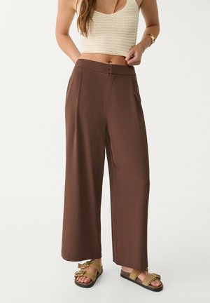 Femme portant un pantalon marron à jambes larges, un crop top en maille crème et des sandales à boucle dorée, debout devant un fond clair uni.