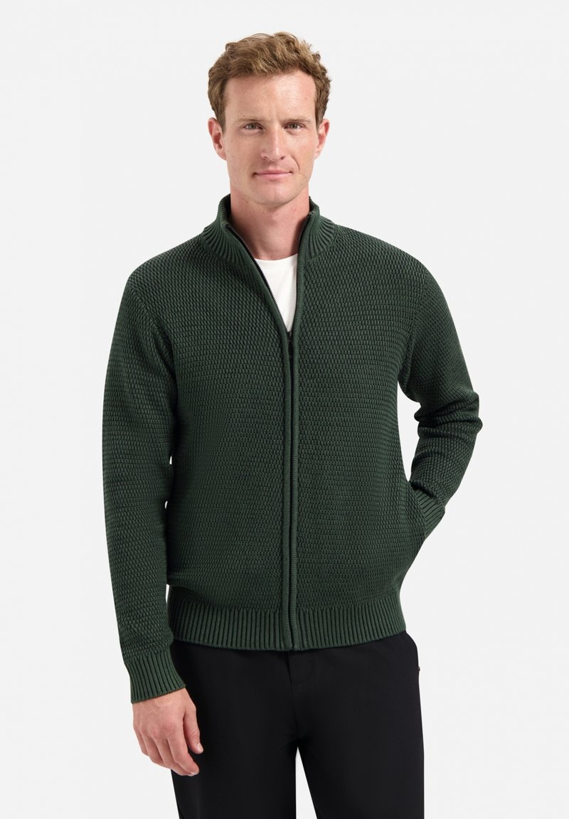 Groene rits-sweater met een textuurpatroon, hoge kraag en geribbelde manchetten en zoom. Draag het over een wit shirt met zwarte broek.
