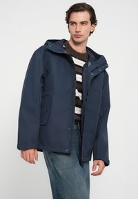 Ναυτικό αδιάβροχο μπουφάν με κουκούλα, κλείσιμο με φερμουάρ και αναδιπλούμενα κουμπιά, φορεμένο πάνω από ένα καφέ και λευκό ριγέ πουλόβερ.