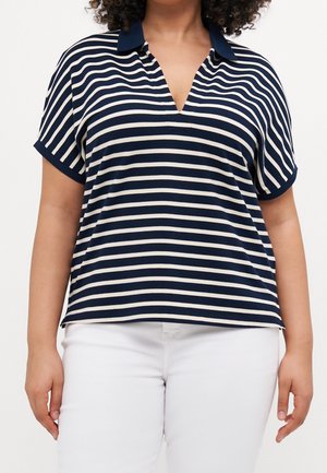 Frau trägt ein kurzärmeliges, marineblau-weiß horizontal gestreiftes Hemd mit Kragen und weiße Hose vor einem einfarbigen Hintergrund.