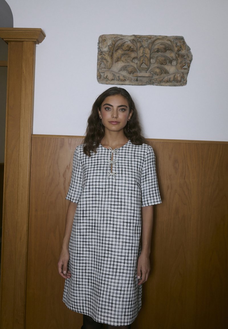 Karo obleko v črno-beli barvi s kratkimi rokavi, okroglim izrezom in detajlom gumbka v obliki ključavnice spredaj. Strukturirano blago.