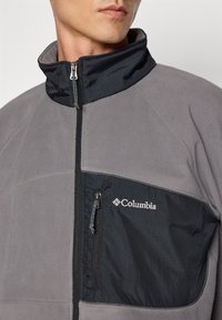 Giacca in pile grigia con pannello in nylon nero e tasca a zip sul petto. Caratterizzata da un collo alto e dal logo Columbia.