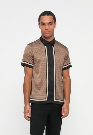 Hombre con una camisa marrón de manga corta y botones, con cuello negro y rayas verticales, combinado con pantalones negros, de pie frente a un fondo blanco.