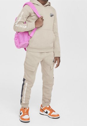 Beige hættetrøje med lomme foran og Nike-logo. Matchende beige joggingbukser med sidelommer og grå striber. Lys pink rygsæk og orange sneakers.