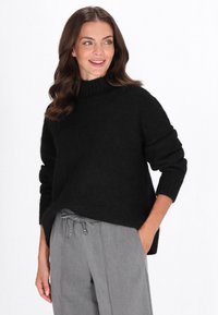 Schwarzer, gestrickter Pullover mit hohem Kragen, lockerer Passform und gerippten Bündchen. Kombiniert mit grauen Hosen mit Kordelzug und Seitentaschen.