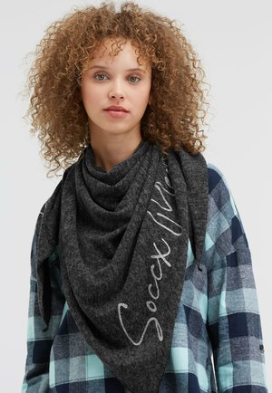 Young person with curly hair wearing a blue checkered shirt and dark gray scarf with "Social vibe" text draped around the neck.