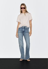 Helles rosa T-Shirt, lockere Passform. Ausgestellte hellblaue Jeans mit einem verblassten Muster. Modell trägt dunkle Sonnenbrillen und Flip-Flops.