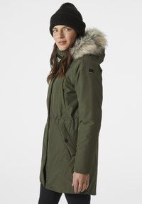 Parka verde oliva con capucha desmontable con pelo, cintura elástica, cierre frontal con cremallera y bolsillos laterales. Lleva un gorro de punto negro.