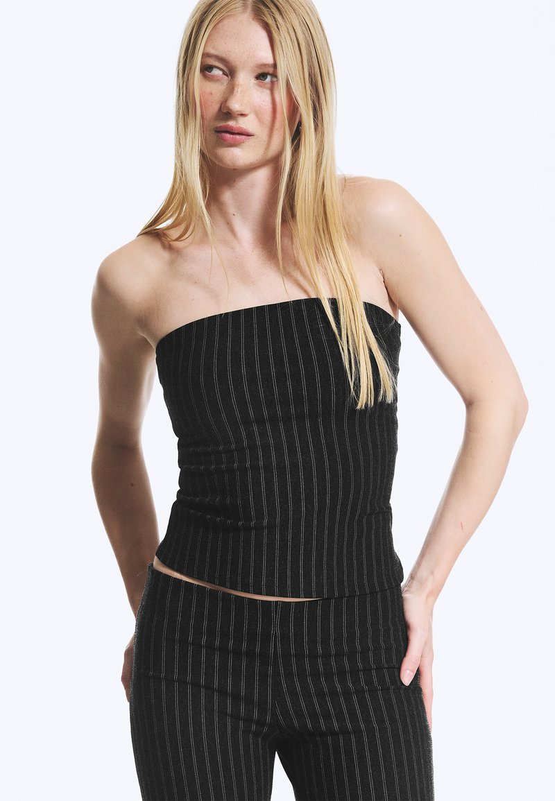 Femme aux longs cheveux blonds portant un haut noir à rayures sans bretelles et un pantalon assorti, debout devant un fond clair et uni.