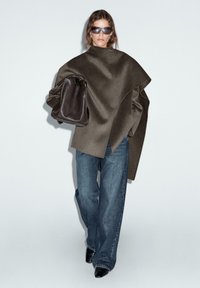 Overmaatse bruine wolblend jas met asymmetrisch ontwerp, gecombineerd met lichtblauwe denim wijdere jeans en zwarte puntige laarzen. Bruin leren handtas.