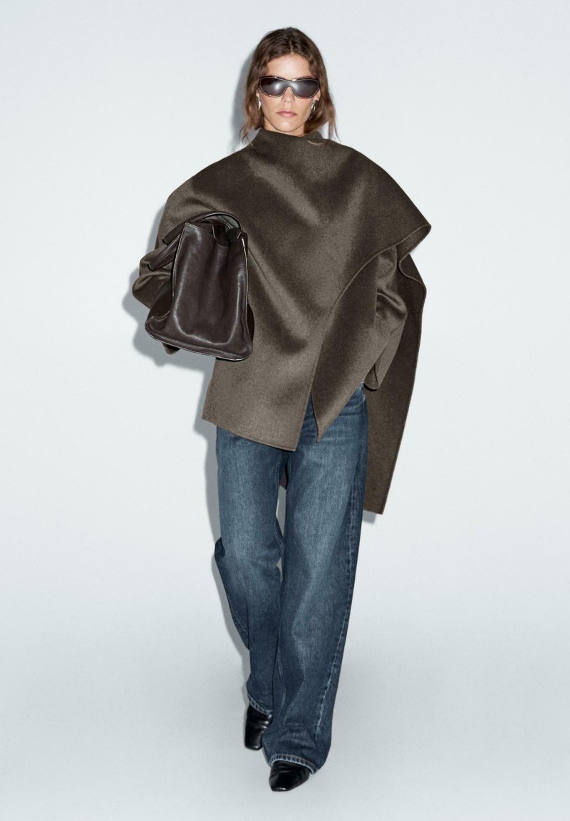 Overmaatse bruine wolblend jas met asymmetrisch ontwerp, gecombineerd met lichtblauwe denim wijdere jeans en zwarte puntige laarzen. Bruin leren handtas.